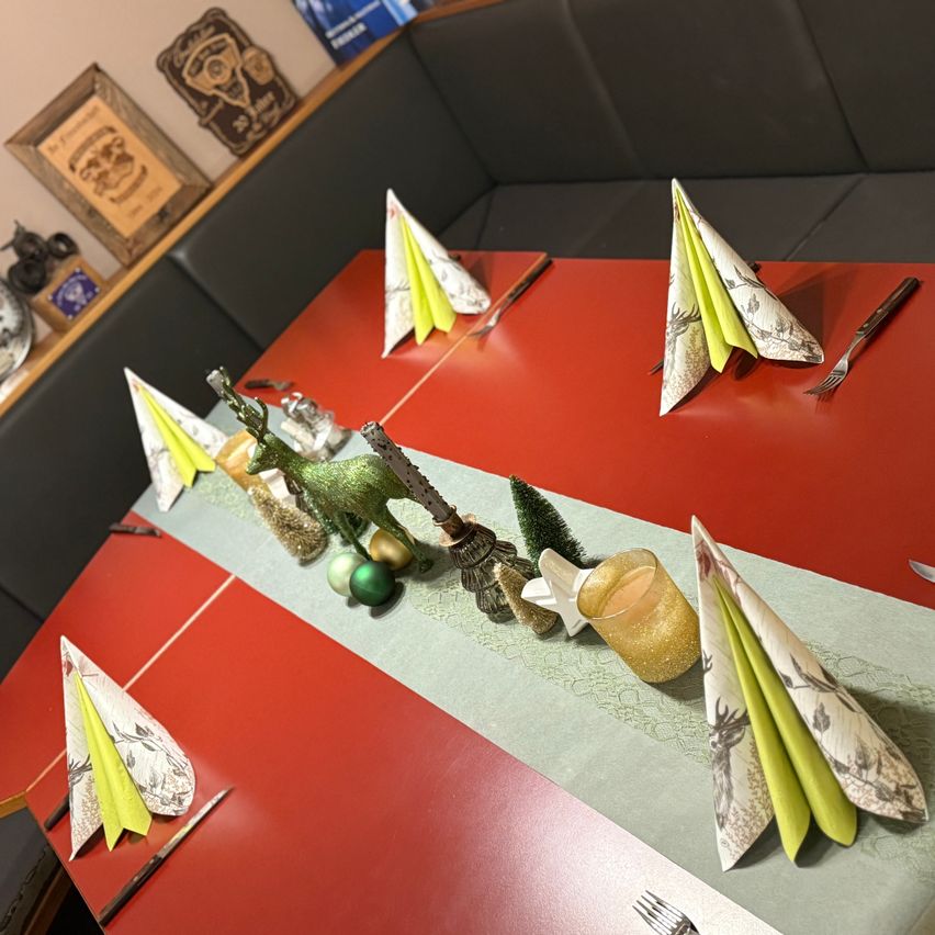 Ein roter Esstisch ist mit Weihnachtsdekorationen wie grünen Bäumen, Ornamenten und in Dreiecke gefalteten Papierservietten gedeckt. Es gibt Messer, Gabeln und eine Kerze auf dem Tisch.