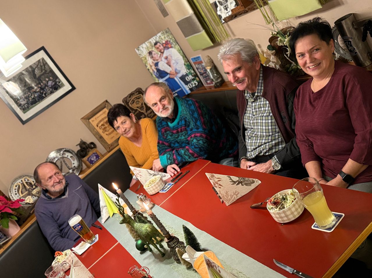 Fünf Menschen sitzen um einen roten Tisch mit brennenden Kerzen und festlichen Dekorationen und lächeln. Der Tisch ist mit Tellern, einer Schüssel und Gläsern gedeckt. Im Hintergrund befinden sich gerahmte Bilder und ein Plakat mit einem Datum.