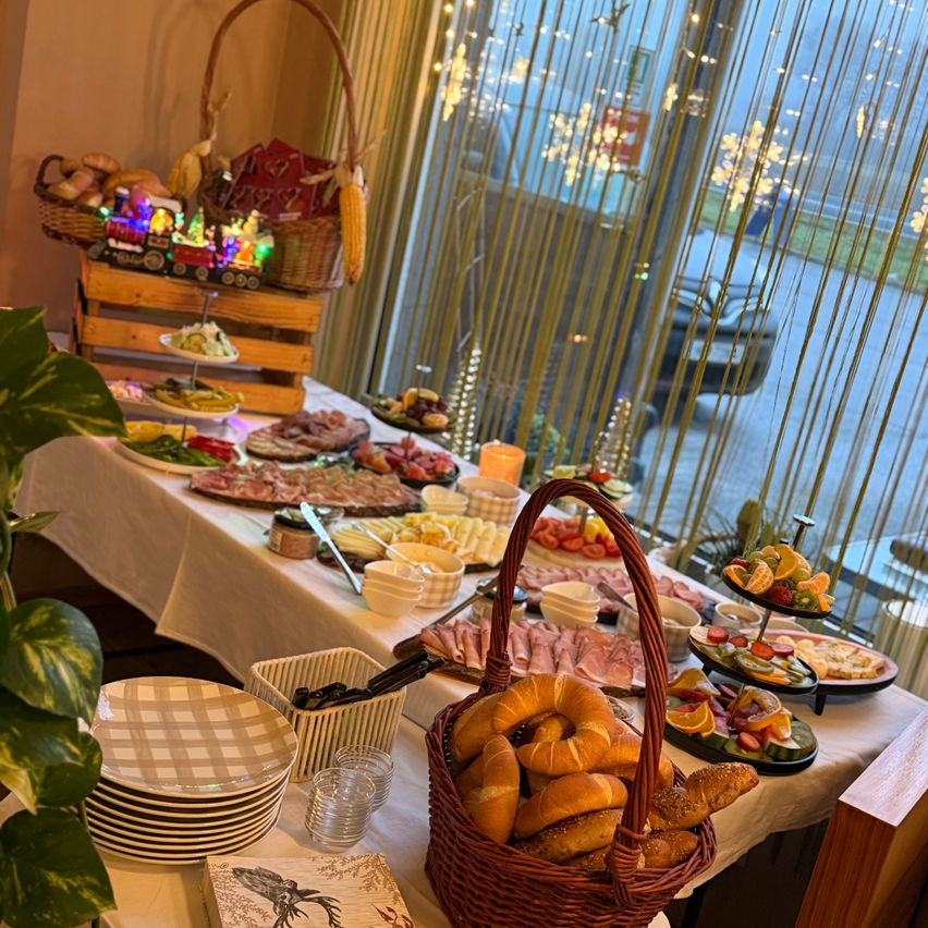 Ein gut gedeckter Tisch mit einer Vielzahl von Speisen auf einem Buffet, einschließlich Brot, Obst, Fleisch und Gemüse. Der Tisch ist mit einer weißen Tischdecke bedeckt.