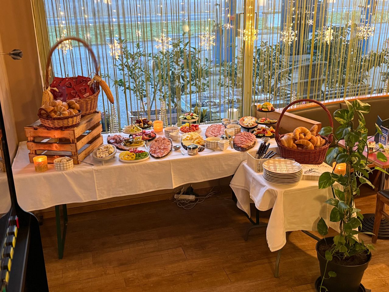 Ein Tisch mit weißer Tischdecke ist mit verschiedenen Lebensmitteln wie Brot, Käse und Gemüse beladen. Zwei Körbe mit Brot befinden sich auf beiden Seiten des Tisches. Eine Pflanze steht auf der rechten Seite.