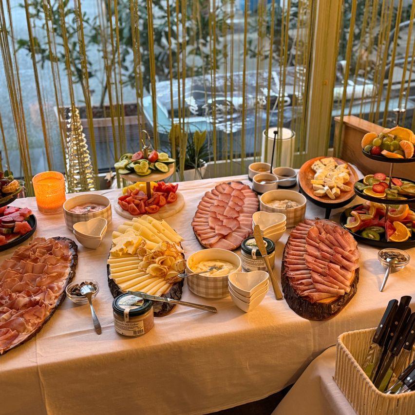 Ein Buffettisch mit verschiedenen Fleischsorten, Käse, Obst und Beilagen. Der Tisch ist mit einem weißen Tuch bedeckt. Eine Kerze und Topfpflanzen im Hintergrund.