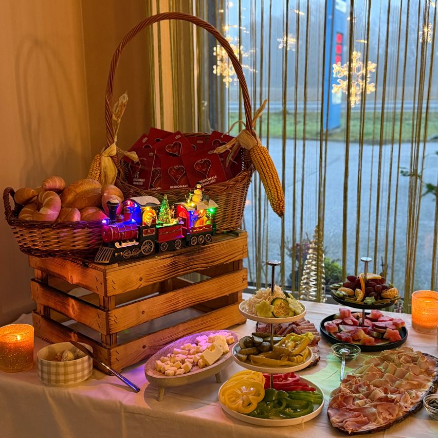 Ein festlicher Tisch mit einem Korb Brot, einem Holzkisten mit einem Zug und verschiedenen Speisenplatten, einschließlich Käse, Obst und Fleisch. Kerzen und Dekorationen verstärken die festliche Stimmung.