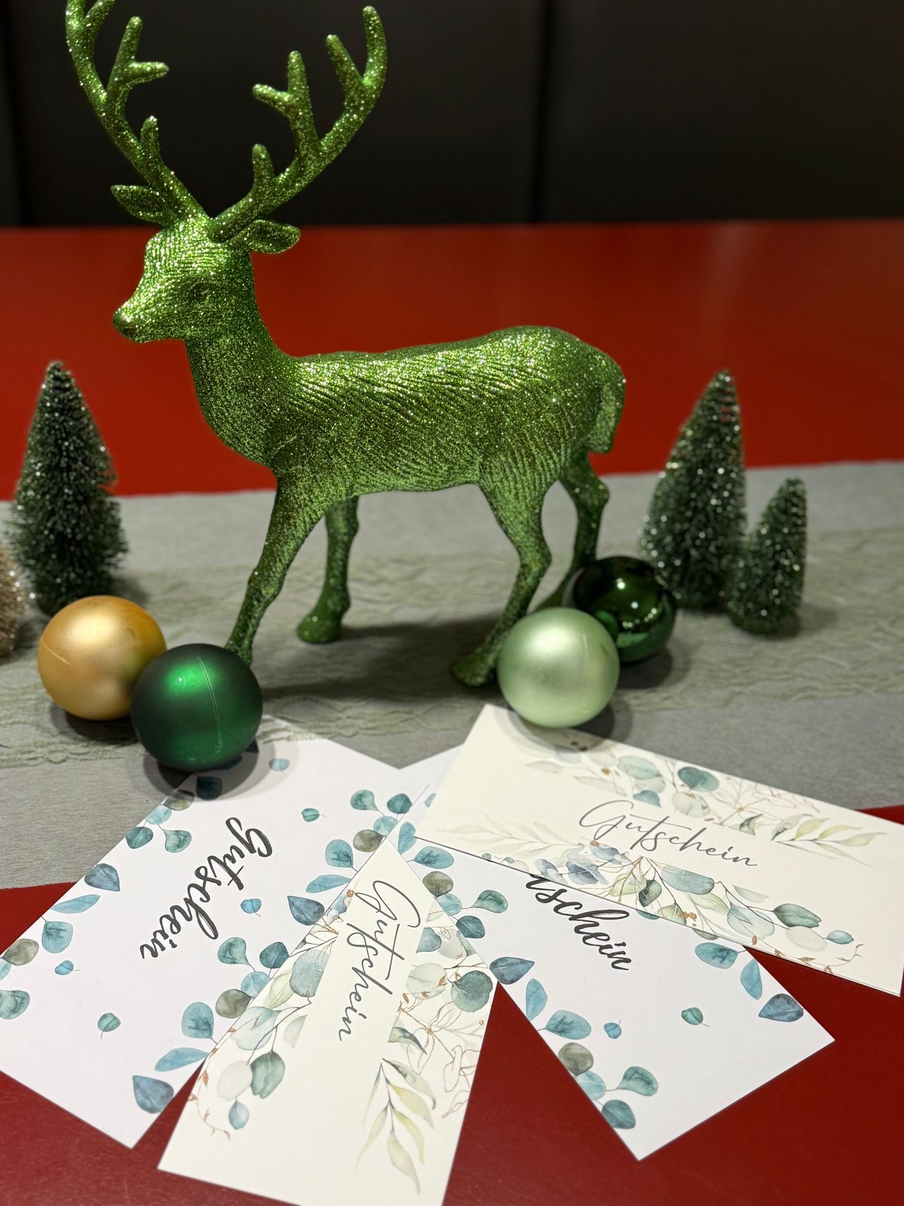 Ein grüner glitzernder Hirsch steht auf einem roten Tisch mit grünen Ornamenten und kleinen Weihnachtsbäumen. Daneben liegen Weihnachtsgeschenkkarten mit 'Christmas' und 'Geschenke' darauf.