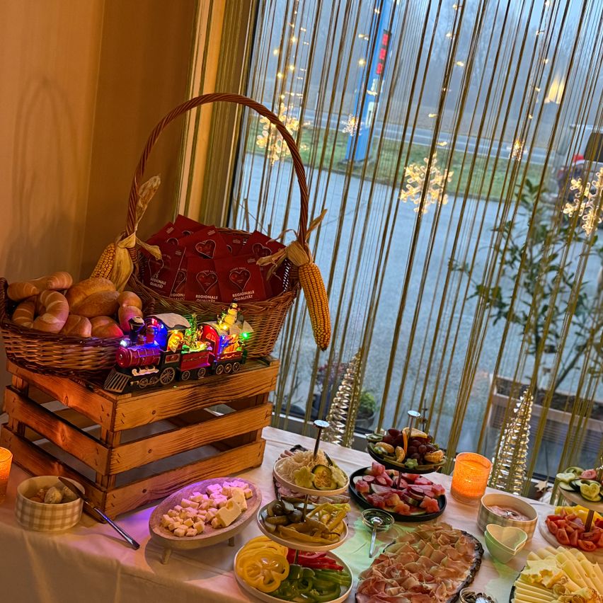 Ein festlicher Buffettisch voller verschiedener Speisen, einschließlich Käse, Fleisch und Gemüse. Ein Korb mit Brot und eine beleuchtete Lokomotive stehen auf einem Holzkisten. Eine beleuchtete Kerze und Weihnachtsdekorationen sind vor dem Fenster zu sehen.