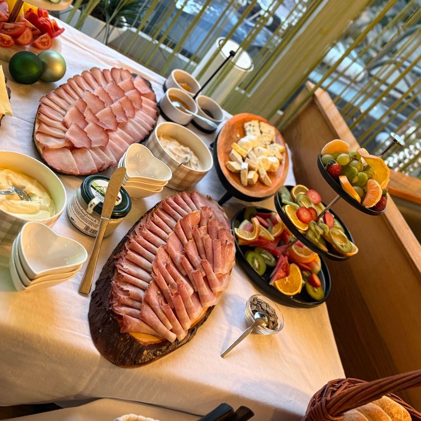 Ein Tisch ist mit einer Vielfalt an Speisen gedeckt, darunter Fleisch, Käse, Obst und Brot. Es gibt zwei große Platten mit geschnittenem Schinken, eine Schüssel Salat und eine Platte mit Käse.