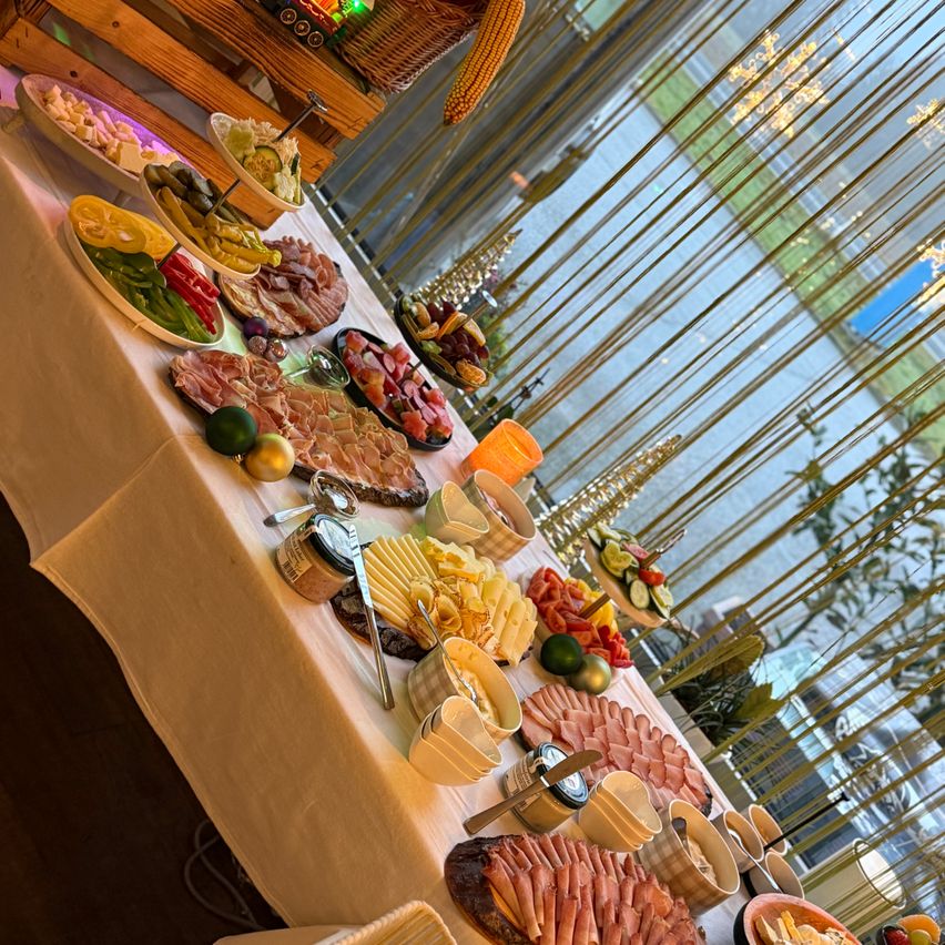 Ein langer Tisch mit weißer Tischdecke ist gefüllt mit verschiedenen Speisen, darunter Fleisch, Käse, Gemüse und Obst. Der Tisch ist mit einer Kerze und einigen festlichen Dekorationen geschmückt. Die Aussicht nach draußen zeigt ein Gewässer mit der Reflexion von Bäumen.