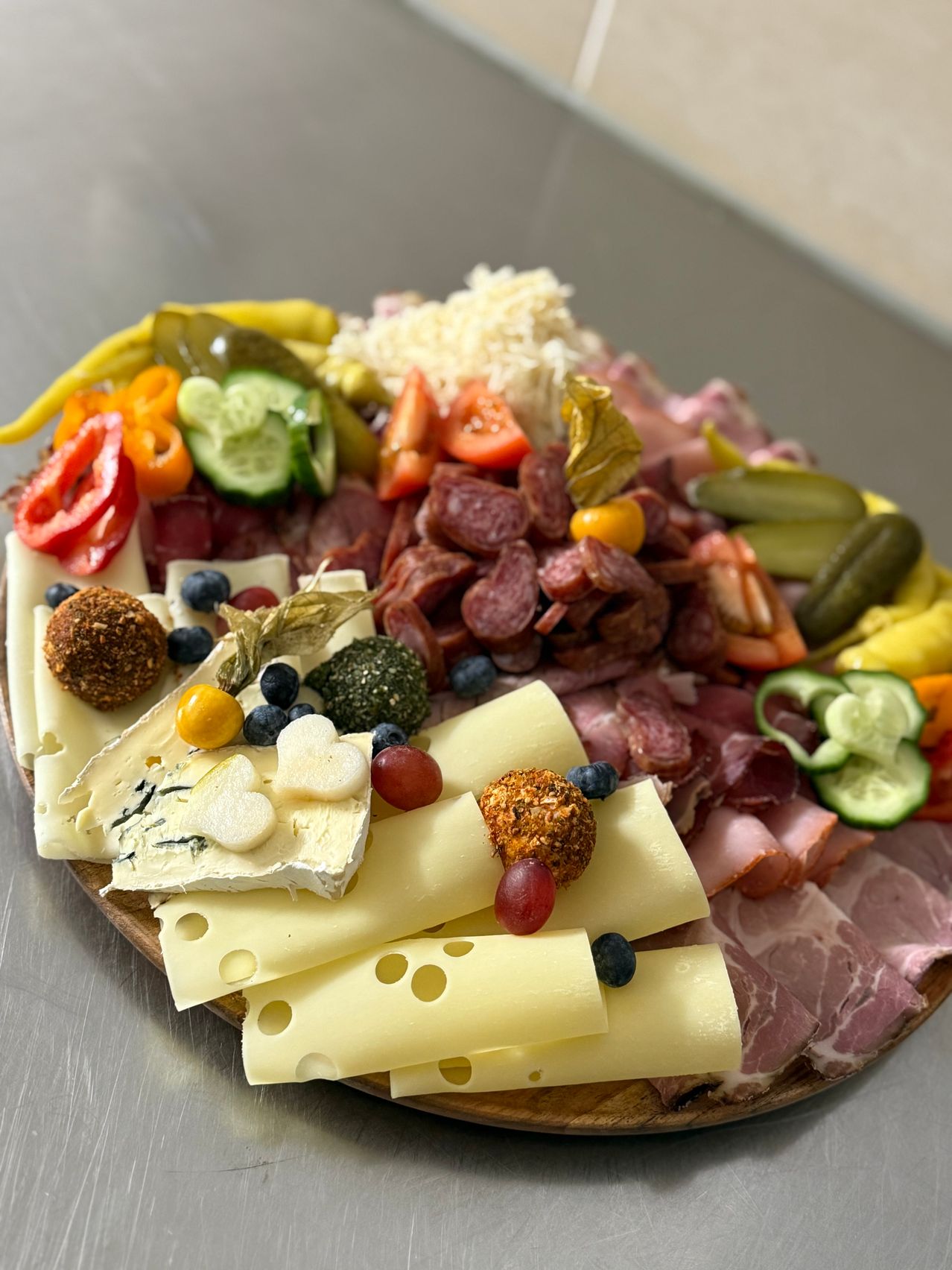 Ein Holzbrett zeigt eine Auswahl an Fleisch, Käse und Gemüse, darunter Salami, Trauben, Blaubeeren und Gurken.