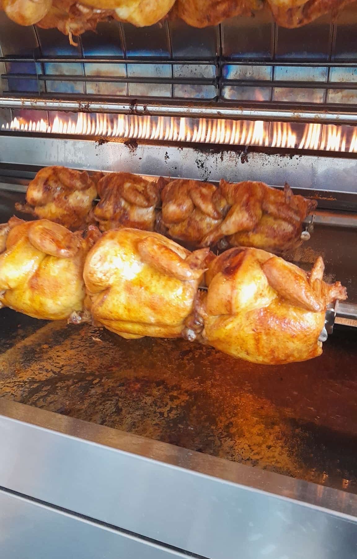 Several roasted chickens on a rotisserie. They are cooked in a large oven with visible flames. The chickens are golden brown with crispy skin.