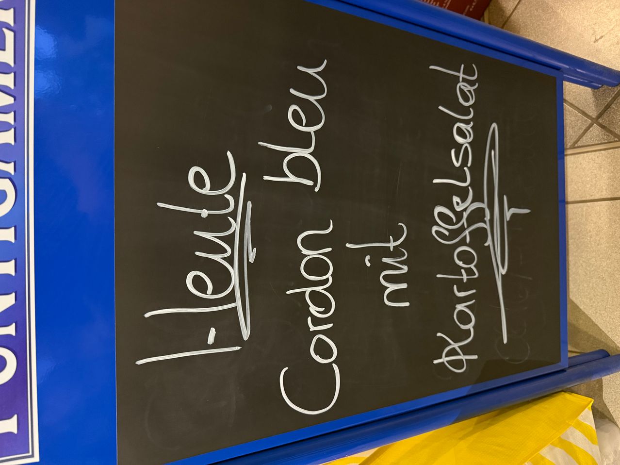 Ein schwarzes Tafelmenü zeigt 'Newell Cordon Bleu', 'Milz' und 'Kartoffelsalat'. Rechts befindet sich eine Unterschrift.