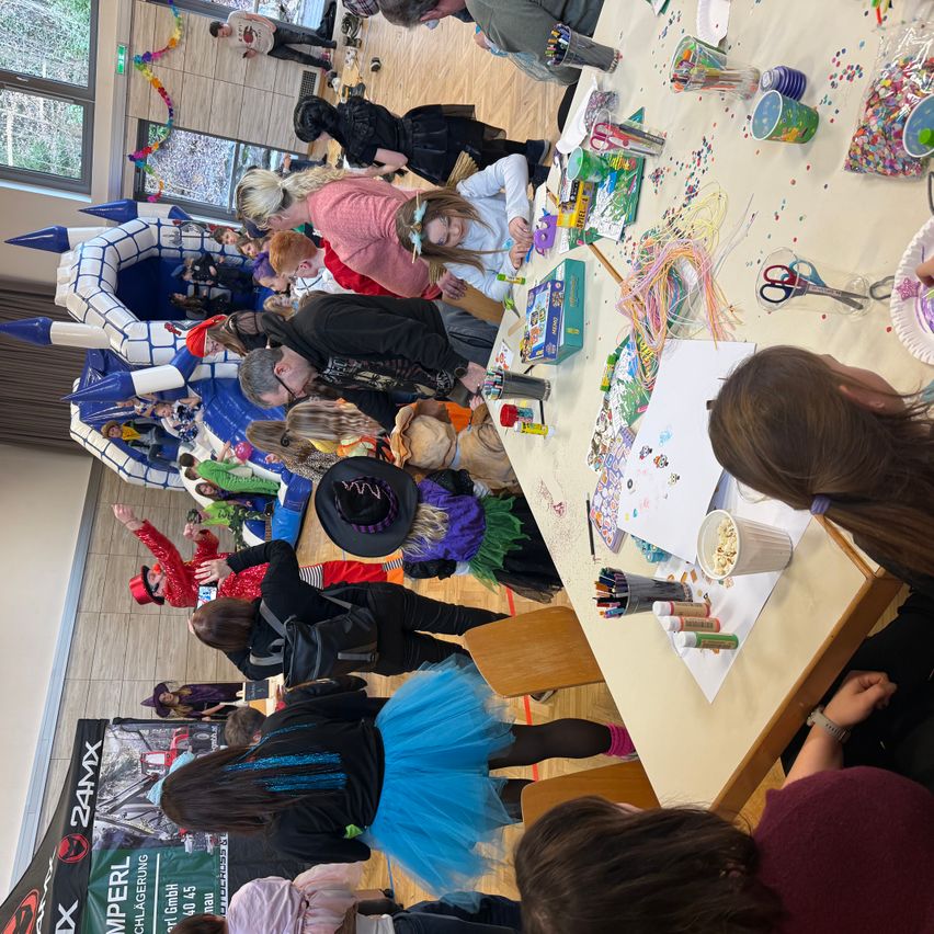 Eine Gruppe von Menschen, darunter Kinder, ist in einem Raum für eine Bastelveranstaltung versammelt. Viele arbeiten an Bastelarbeiten, während andere in der Schlange in der Nähe eines bunten aufblasbaren Schlosses warten.