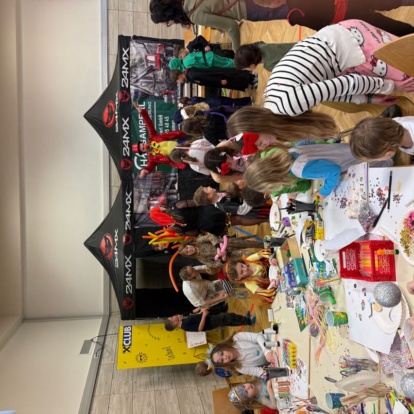 Mehrere Kinder sind um einen Tisch mit verschiedenen Bastelmaterialien versammelt. Sie sind in einer Bastelaktivität verwickelt. Ein Banner im Hintergrund trägt die Aufschrift 24MX.