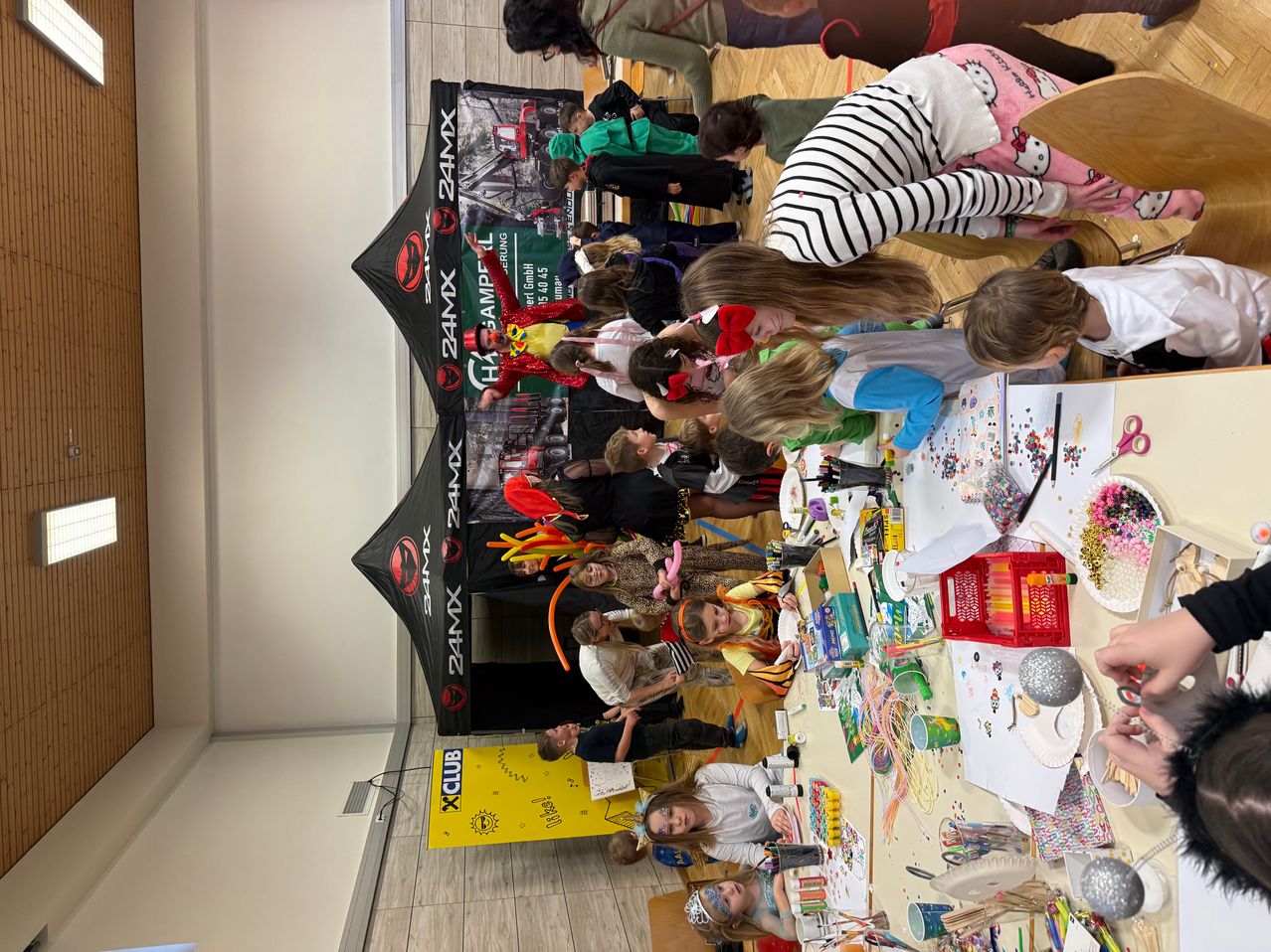 Mehrere Kinder sind um einen Tisch mit verschiedenen Bastelmaterialien versammelt. Sie sind in einer Bastelaktivität verwickelt. Ein Banner im Hintergrund trägt die Aufschrift 24MX.