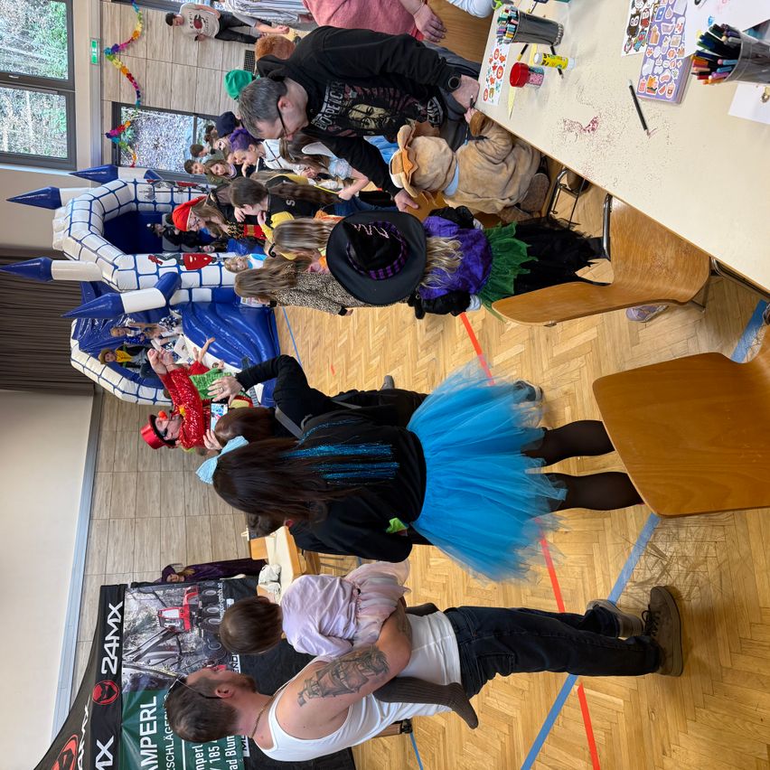 Eine große Gruppe von Menschen, einschließlich Erwachsener und Kinder, ist drinnen versammelt. Sie nehmen an einer Veranstaltung teil, einige sind in Kostümen gekleidet. Ein Tisch mit Bastelmaterial steht auf der rechten Seite. Ein aufblasbarer Turm befindet sich im Hintergrund.