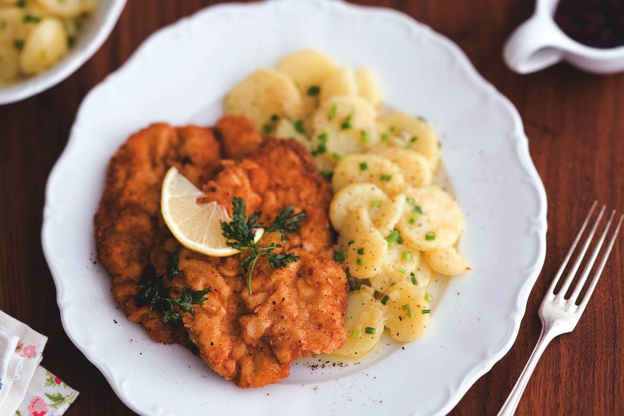 Auf einem weißen Teller liegt ein goldbraunes Gericht aus panierter Chickenschnitzel mit einer Zitronenscheibe, garniert mit Petersilie. Daneben befindet sich eine Portion cremiger Kartoffelpüree mit Schnittlauch.