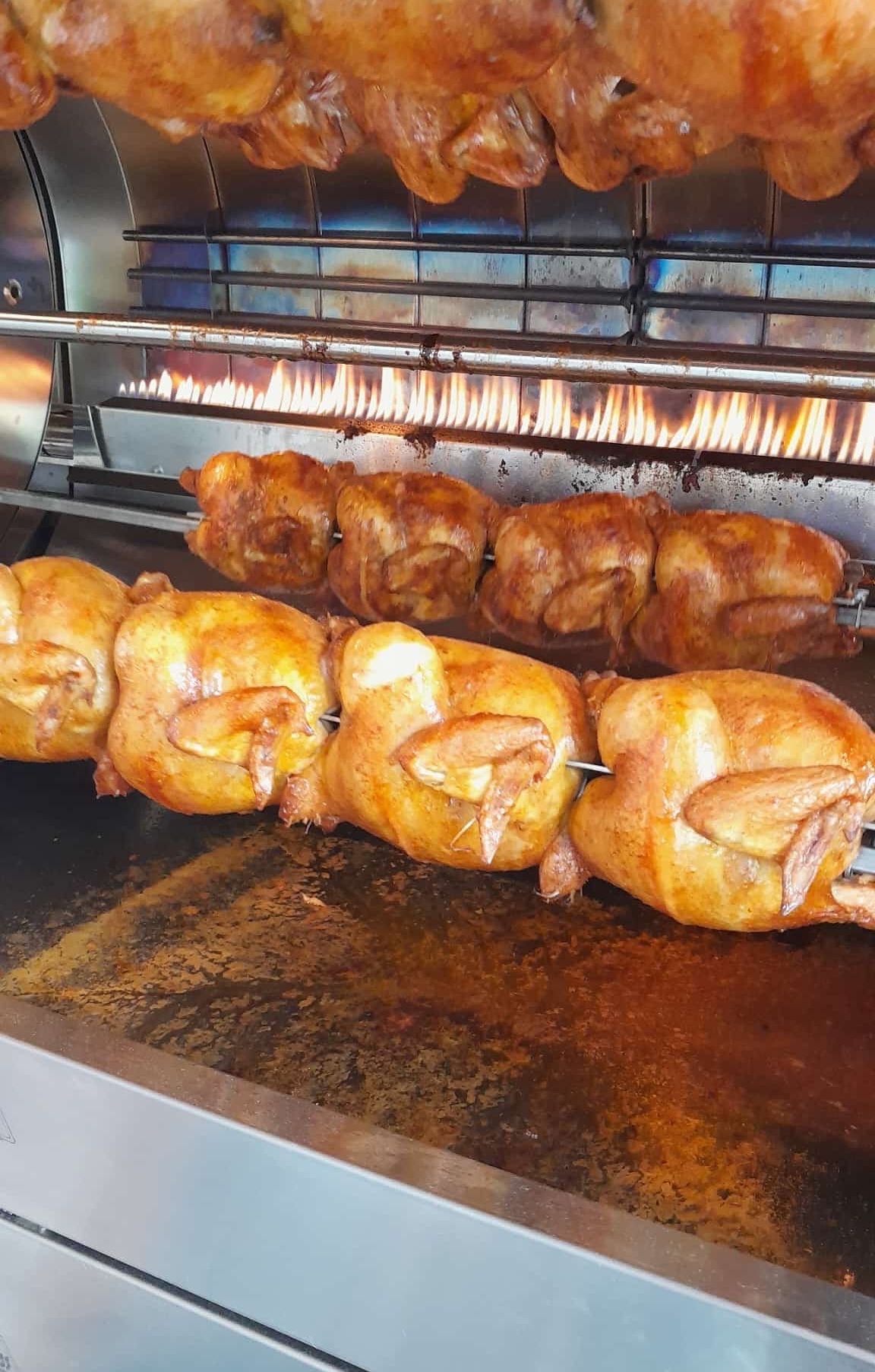 Mehrere ganze Hühner werden auf einem Grill geröstet. Der Grill ist mit Flammen gefüllt, und die Hühner haben eine goldbraune Farbe.