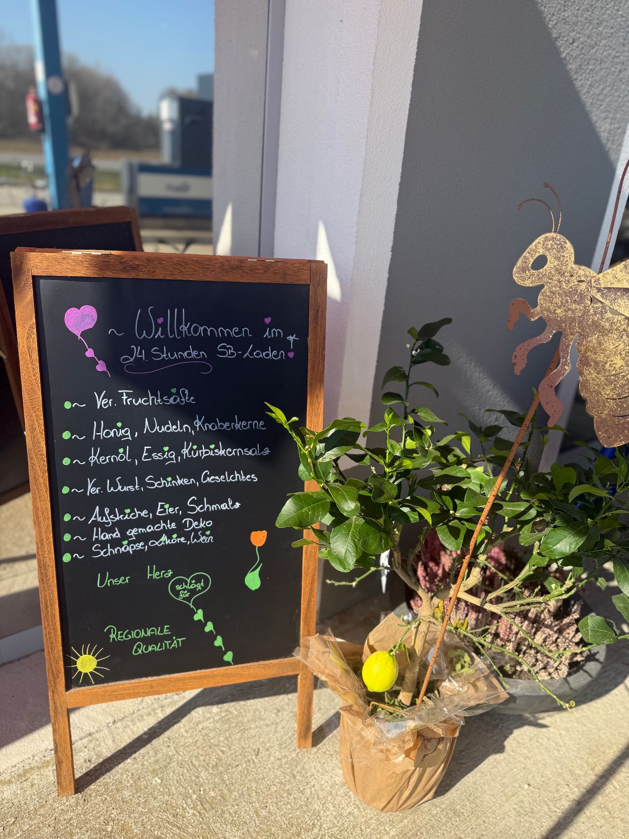 Ein Kreidetafel mit Willkommensnachricht und Speisekarte ist im Freien ausgestellt. Es listet Artikel wie frische Säfte, Honig und mehr auf. Eine Bienenstatue und eine Zitronenpflanze sind in der Nähe.
