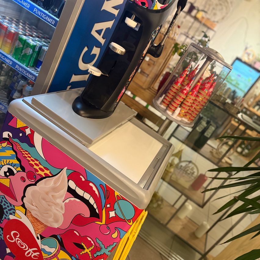 Ein farbenfroher Eisspender mit einer Kugel Eis darauf, neben einem Kühler voller Eisbecher.