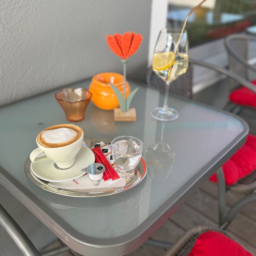 Auf einem Tisch stehen eine Kaffeetasse mit Schaum, eine Untertasse und ein Glas Wasser, begleitet von einem Weinglas mit Zitronenscheibe und einer Blumenarrangement.