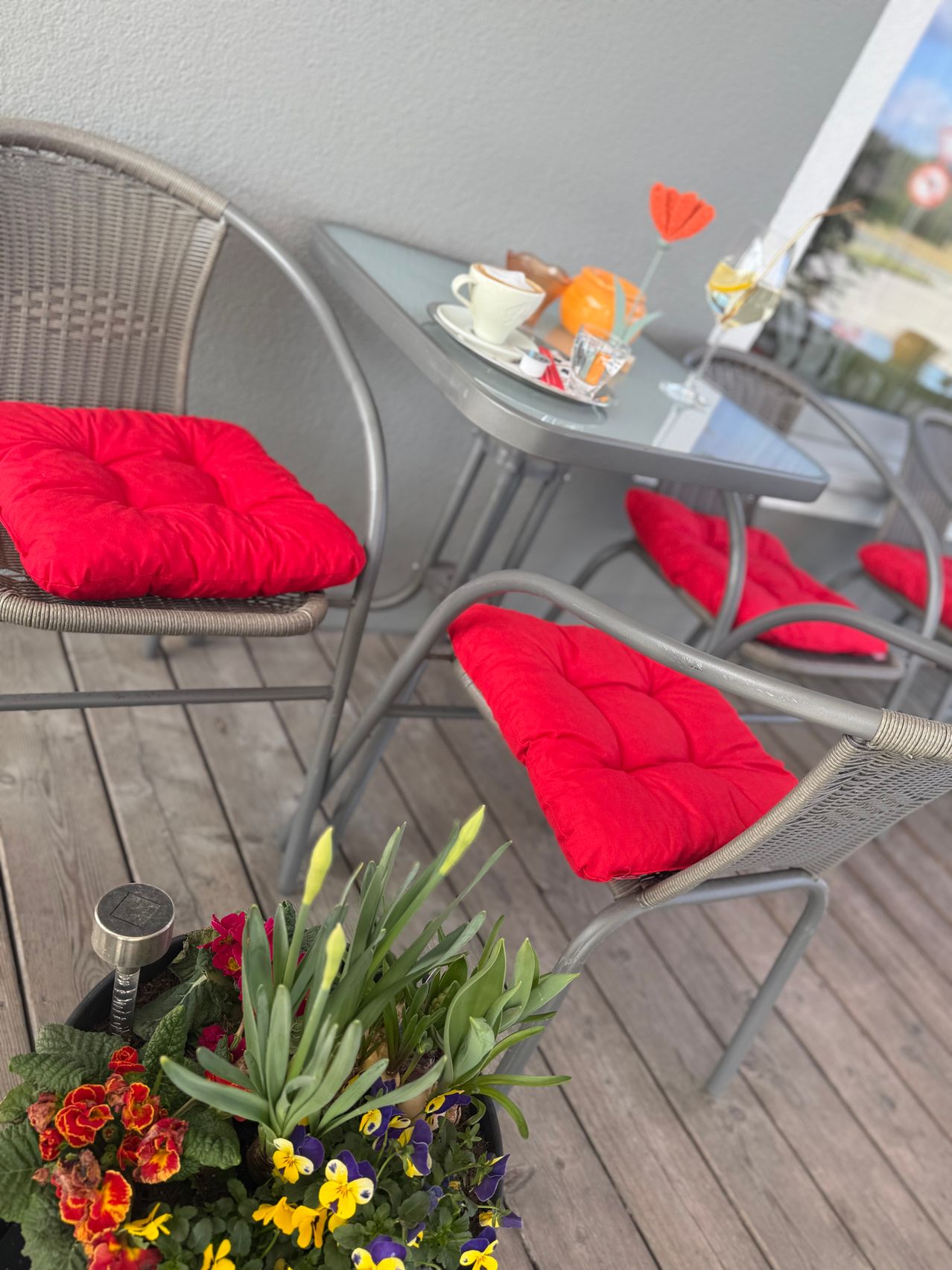 Eine Terrassengarnitur mit vier Stühlen um einen kleinen Tisch, jeder Stuhl hat einen roten Kissenbezug. Auf dem Tisch stehen eine weiße Tasse, ein Untertasse, ein Weinglas und Blumen.