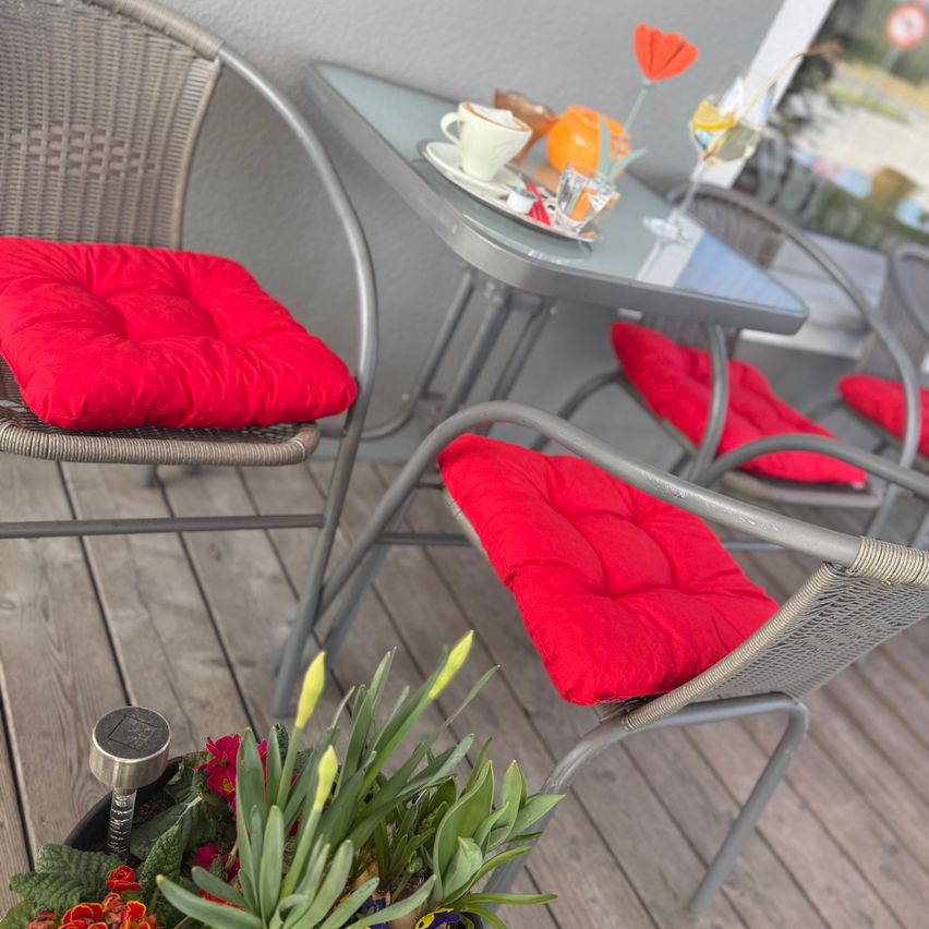 Eine Terrassengarnitur mit vier Stühlen um einen kleinen Tisch, jeder Stuhl hat einen roten Kissenbezug. Auf dem Tisch stehen eine weiße Tasse, ein Untertasse, ein Weinglas und Blumen.