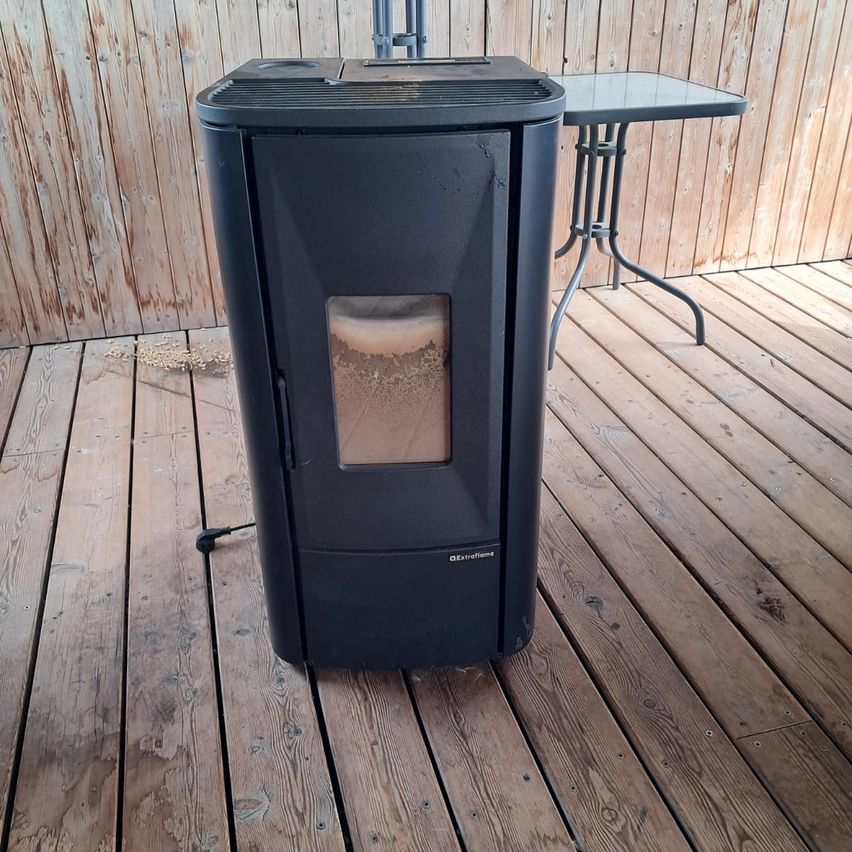 Ein schwarzer Holzofen steht auf einer Holzterrasse, neben einem Glastisch und einem Metallständer. Der Ofen hat ein Glasfenster, durch das ein Feuer im Inneren zu sehen ist.