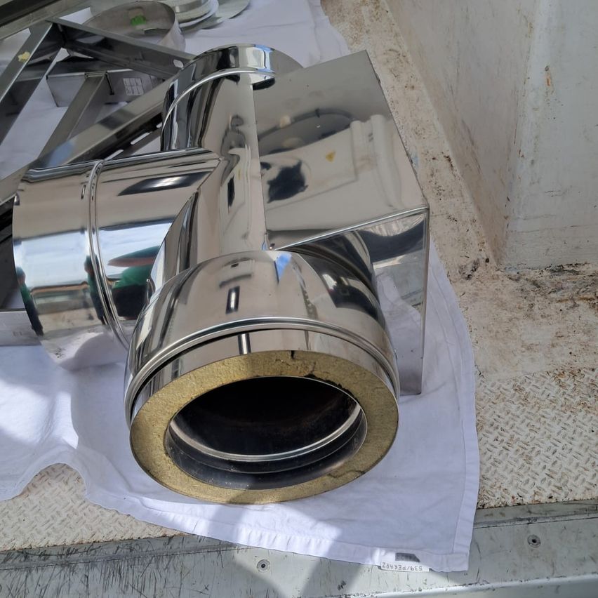 Nahaufnahme eines Schornsteinteils aus Edelstahl mit goldfarbener Basis auf einem weißen Tuch, mit anderen Metallgegenständen im Hintergrund.