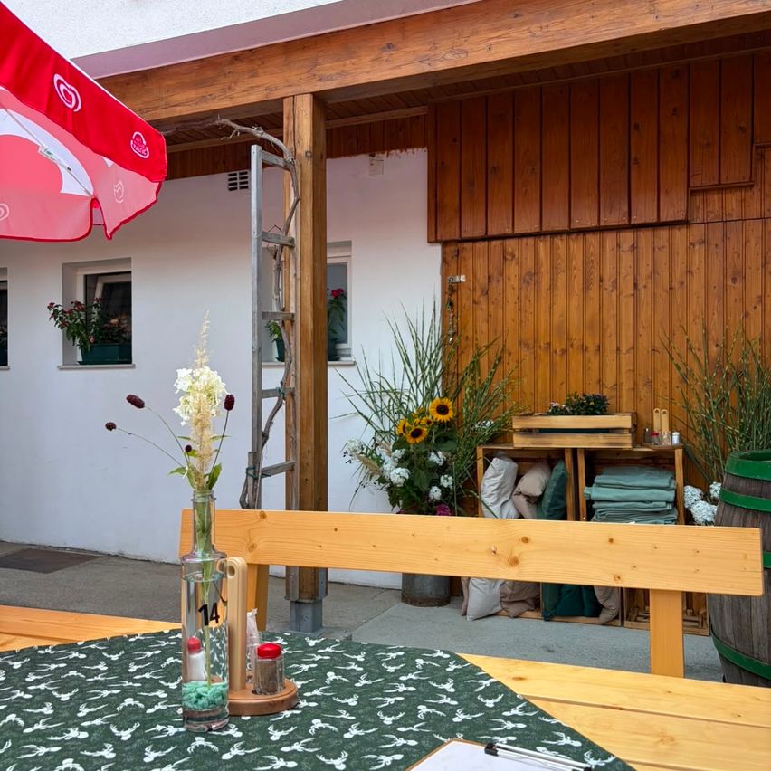 Ein hölzerner Tisch mit einer Blumenarrangement und einem Nummer 14 Schild steht vor einem rustikalen Haus mit einem roten Schirm und einem Holzfass.