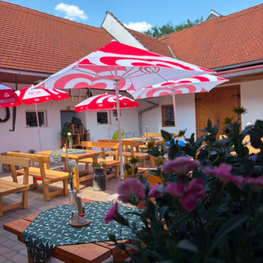 Eine Terrasse eines Restaurants mit Holztischen und -stühlen, roten und weißen Sonnenschirmen und einer Blumendekoration auf einem Tisch.