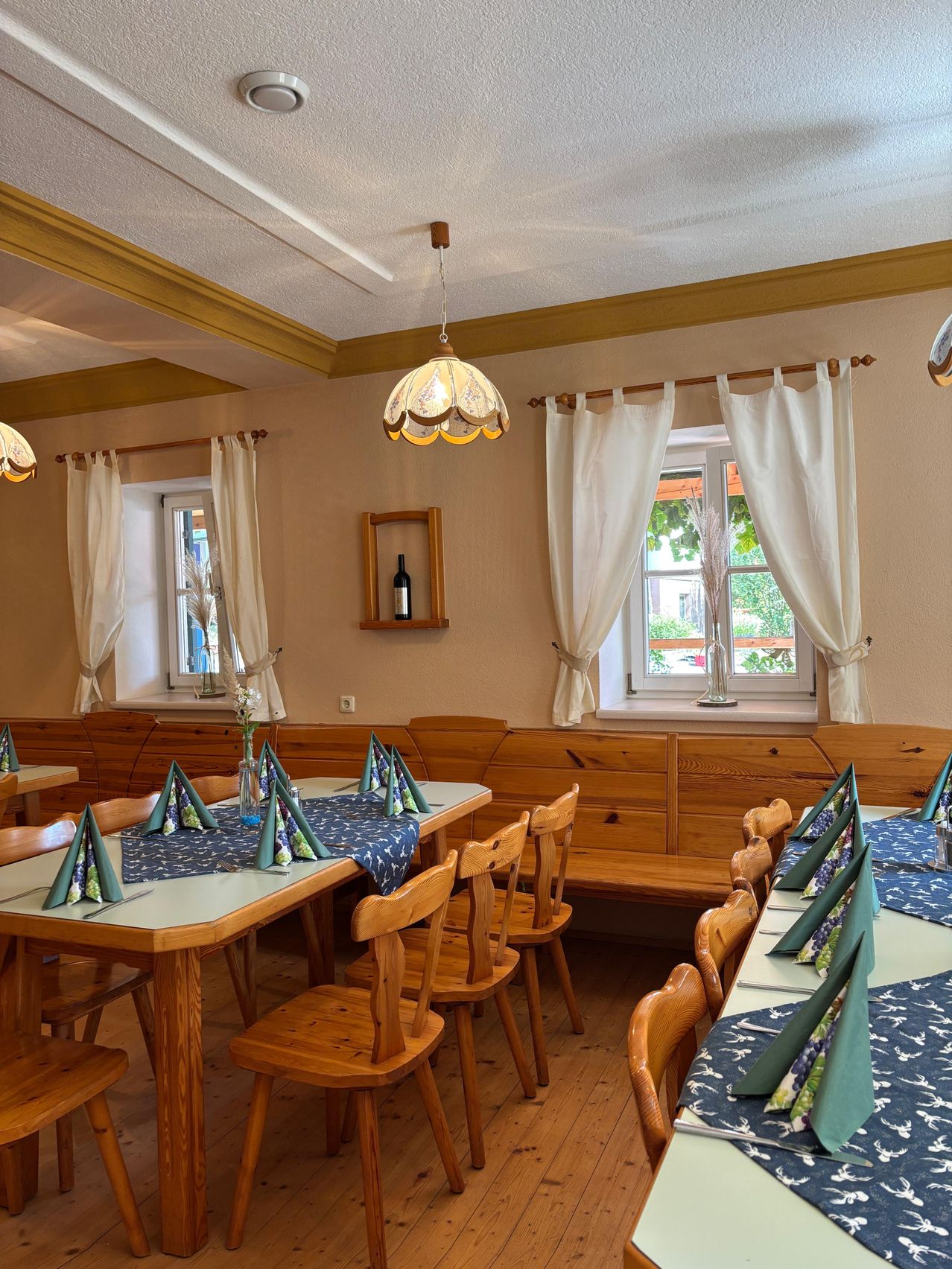 Ein Innenbereich zum Essen mit Holztischen und Stühlen, weißen Vorhängen und einer Flasche auf einem Regal. Der Raum hat ein warmes, rustikales Ambiente.
