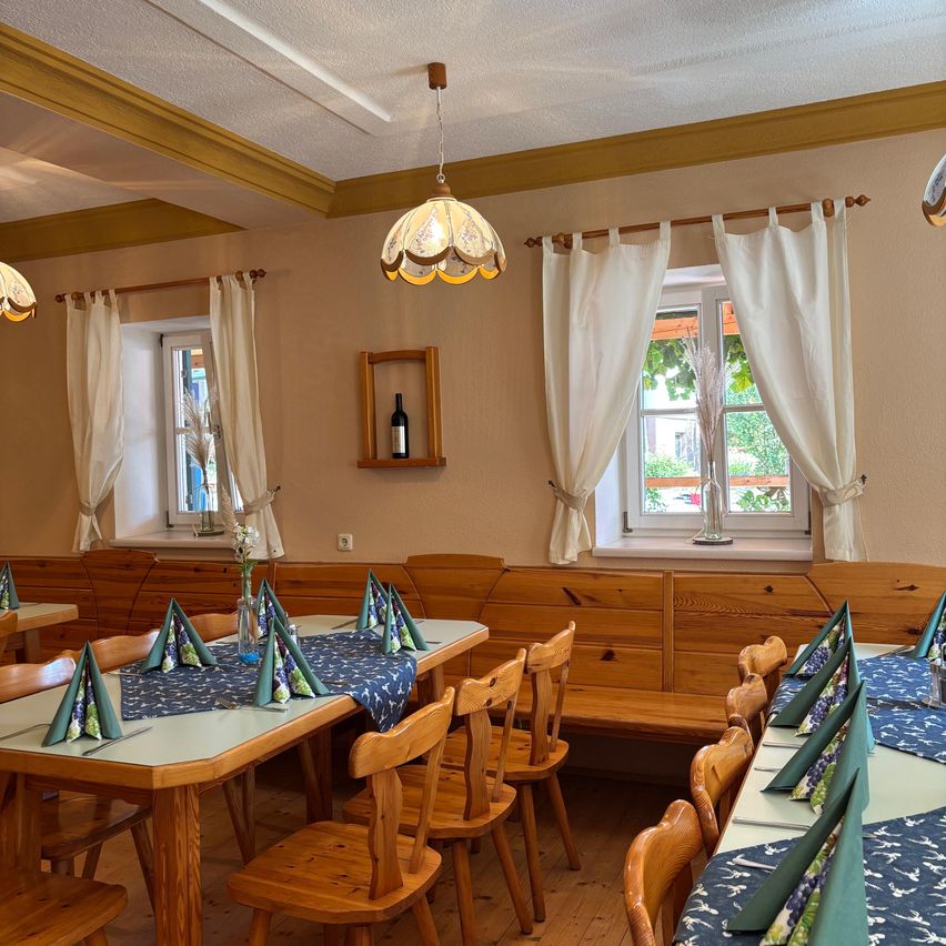 Ein Innenbereich zum Essen mit Holztischen und Stühlen, weißen Vorhängen und einer Flasche auf einem Regal. Der Raum hat ein warmes, rustikales Ambiente.