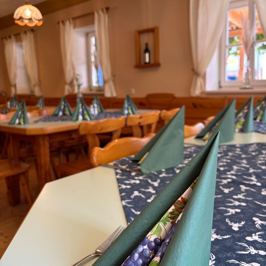 Ein Esszimmer mit mehreren Tischen, die mit blauen und grünen Servietten, Gabeln und Messern gedeckt sind. Der Raum hat Holzmöbel, weiße Vorhänge und ein Holzregal mit einer Flasche.