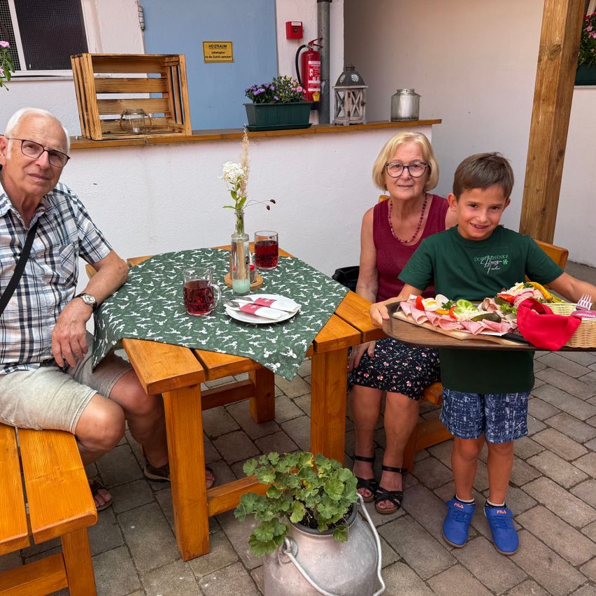 Eine dreiköpfige Familie sitzt an einem Tisch im Garten, ein Junge hält eine Tablett mit Essen. Der Tisch ist mit Getränken und einer Blumenvase bestückt.