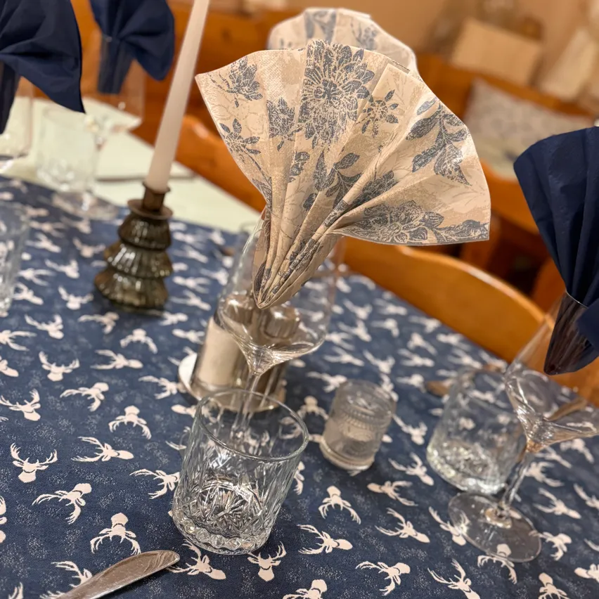 Ein Tisch für eine Mahlzeit mit einer blauen Tischdecke mit Hirschdesigns, einer gefalteten Serviette, einer Kerze, Weingläsern und Besteck.