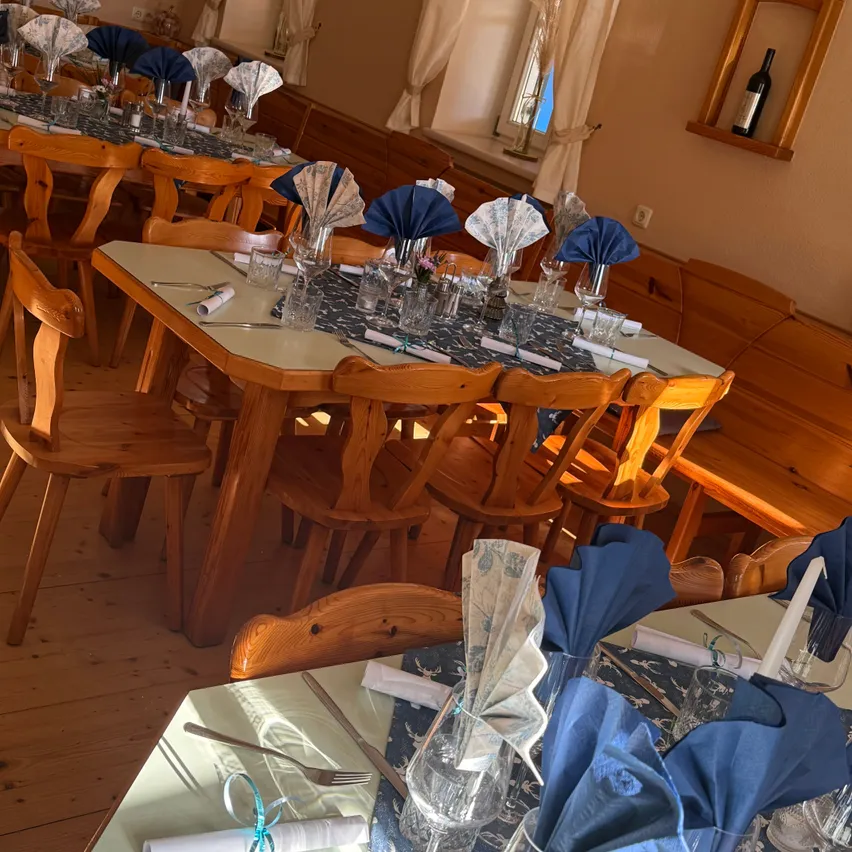 Ein Restaurant-Essbereich mit mehreren Tischen, die für eine Mahlzeit vorbereitet sind. Jeder Tisch hat ein blaues Tuch, ein Weinglas und Besteck. Der Raum hat Holzmöbel und eine warme, einladende Atmosphäre.