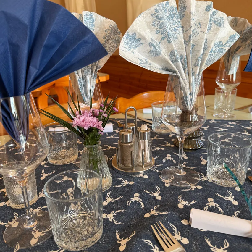 Ein Tisch mit einer blauen Tischdecke mit Hirschdesigns, Weingläsern, einer Vase mit rosa Blumen und gefalteten Servietten.