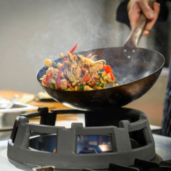 Eine Person kocht ein farbenfrohes Wok-Gemüsegericht über einem Gasbrenner, wobei Dampf aus der Pfanne aufsteigt.