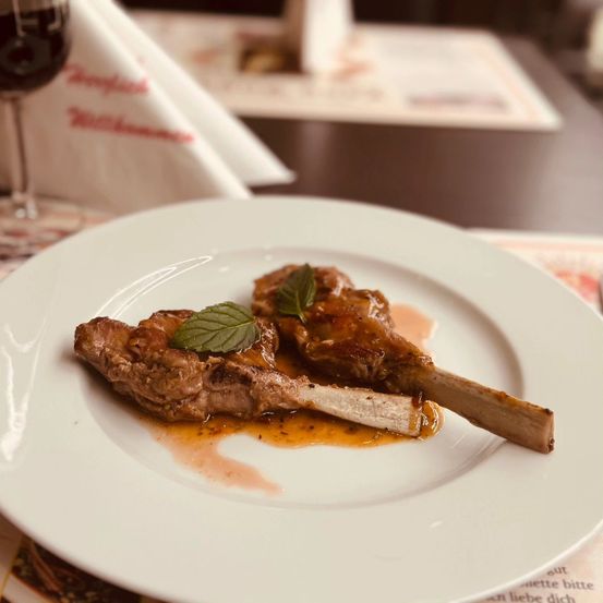 Ein weißer Teller enthält zwei Stücke gebratenes Lamm mit Sauce, garniert mit Minzblättern. Das Ambiente ist elegant, mit einem Weinglas und einem gefalteten Serviettentuch auf dem Tisch.