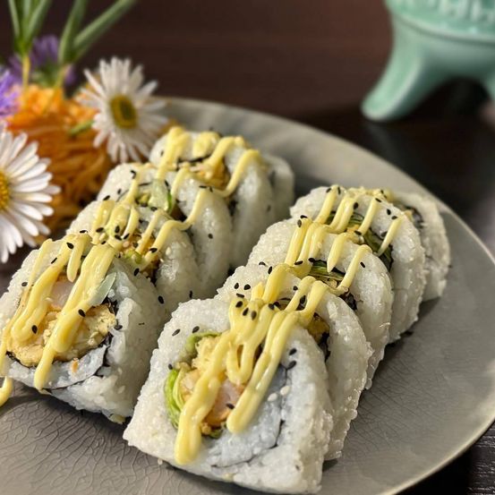 Ein Teller mit Sushi-Rollen, garniert mit weißer Sauce und Sesamkörnern, neben einer Ansammlung von geriebenen Karotten und Blumen.