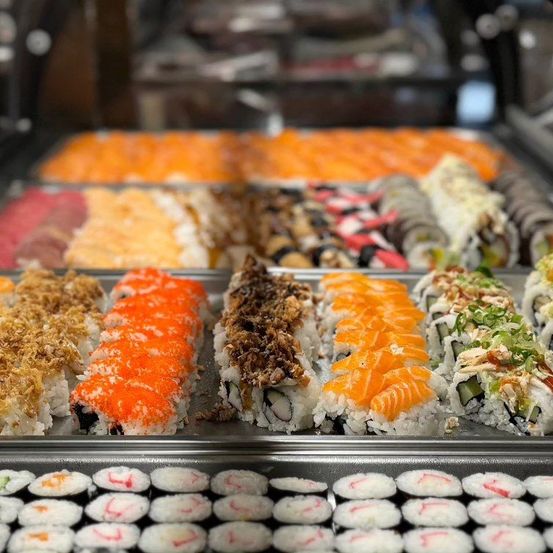 Eine Vitrine mit einer Auswahl an Sushi-Rollen, mit verschiedenen Toppings und Farben, auf silbernen Tabletts in einem Geschäft.