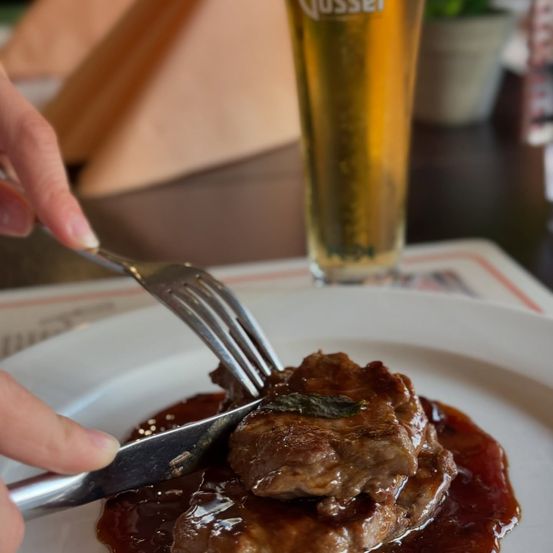 Eine Hand hält eine Gabel und ein Messer, die ein Stück Steak schneiden, das mit Sauce bedeckt ist. Ein Glas Bier und eine Topfpflanze sind im Hintergrund.