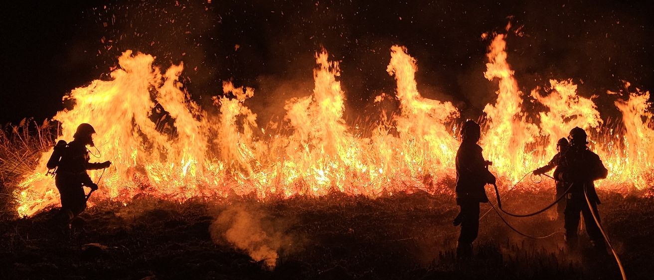 Zwei Feuerwehrleute in Schutzkleidung bekämpfen in der Nacht ein Flächenfeuer. Die Flammen sind intensiv und Funken fliegen in die Luft.