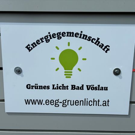 Ein weißes Schild auf einer grauen Oberfläche mit einem grünen Glühbirnensymbol zeigt 'Energiegemeinschaft Grünes Licht Bad Vöslau' und 'www.eeg-gruenlicht.at'.