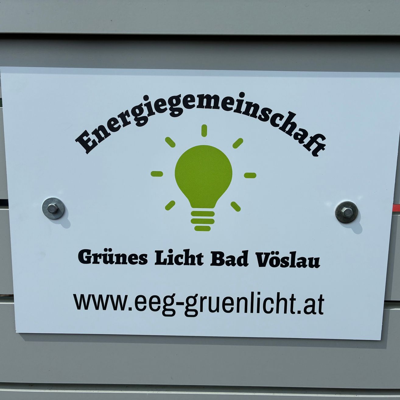 Ein weißes Schild auf einer grauen Oberfläche mit einem grünen Glühbirnensymbol zeigt 'Energiegemeinschaft Grünes Licht Bad Vöslau' und 'www.eeg-gruenlicht.at'.