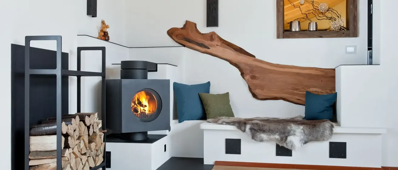 Eine gemütliche Innenfeuerstelle mit Bank, Kissen und Brennholz neben einer Wand mit einem Holzast und einem Regal.