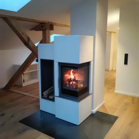 Ein Kamin in einer Ecke eines Raumes mit Holzboden und Balken. Das Feuer brennt mit Holz darin.