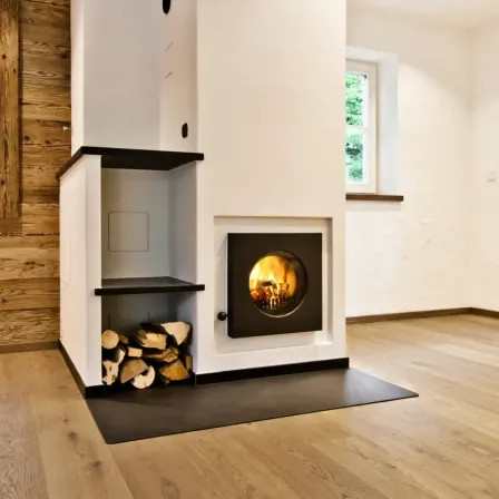 Ein moderner Kamin mit einem brennenden Feuer darin, umgeben von einem Stapel Brennholz. Er steht in einem Raum mit Holzwänden und -böden, mit einem Fenster in der Nähe.