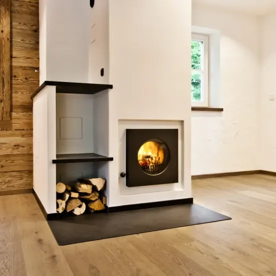 Ein moderner Kamin mit einem brennenden Feuer darin, umgeben von einem Stapel Brennholz. Er steht in einem Raum mit Holzwänden und -böden, mit einem Fenster in der Nähe.