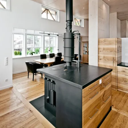 Eine moderne Küche mit Holzboden, einem großen Herd und einem Esstisch. Es gibt Fenster, einen Topf mit Pflanzen und einen Holzschrank.