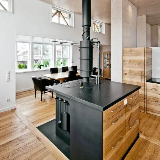 Eine moderne Küche mit Holzboden, einem großen Herd und einem Esstisch. Es gibt Fenster, einen Topf mit Pflanzen und einen Holzschrank.