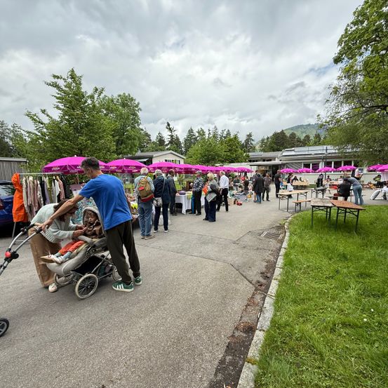 Ein Freiluftmarkt unter bewölktem Himmel mit verschiedenen Ständen, Menschen und Tischen mit rosa Schirmen. Ein Mann schiebt einen Kinderwagen mit einem Baby und einer Frau in der Nähe.